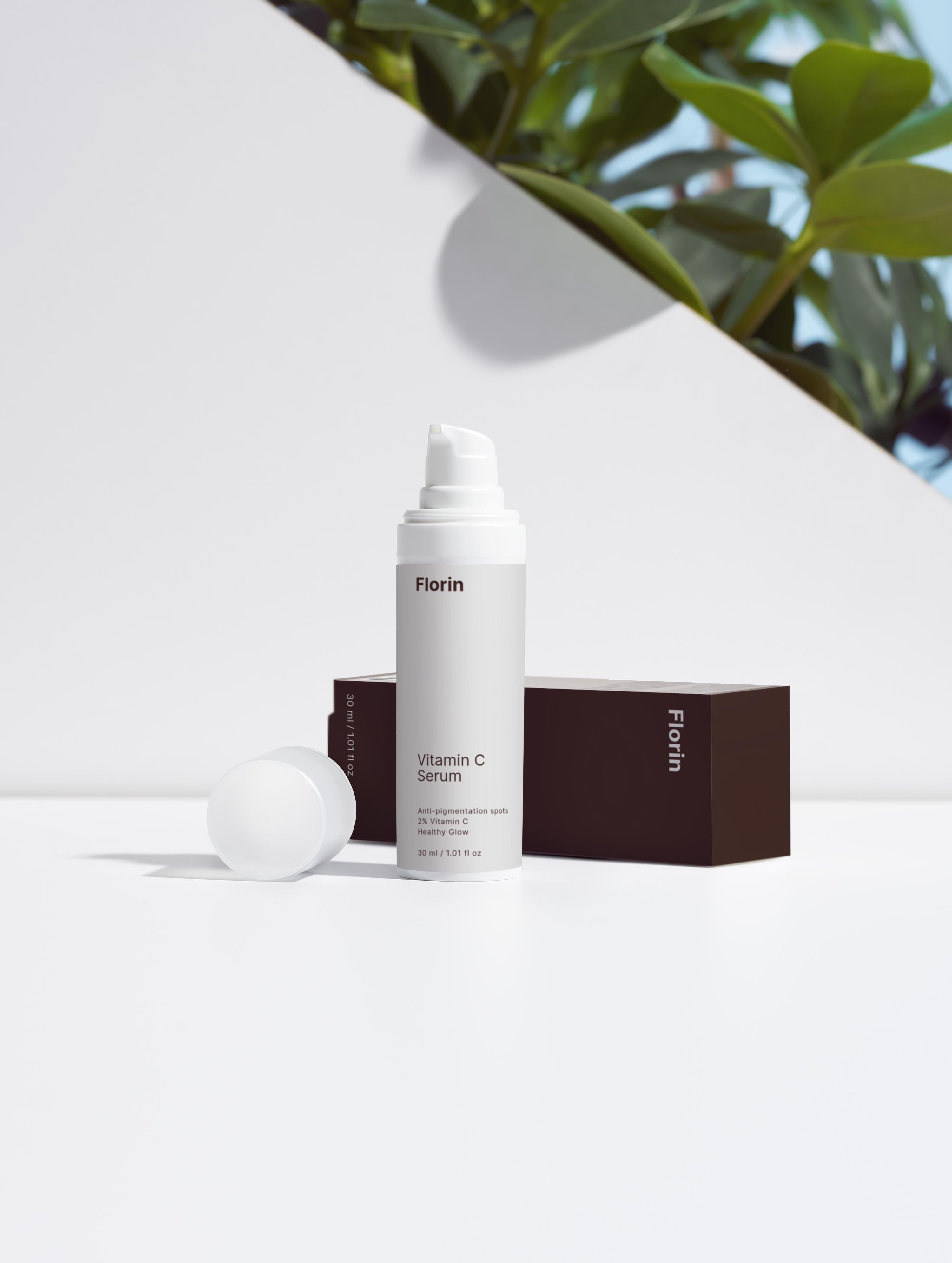 Vitamin C serum bottle with packaging on a white surface with green leaves in the background